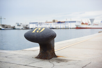 Black marine bollard in harbor with blurred background with ships and industry. - obrazy, fototapety, plakaty