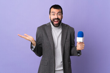 Adult reporter man with beard holding a microphone over isolated purple background with shocked...