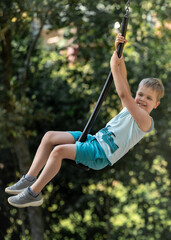 Cute boy dreamily swinging on a rope swing seat against a background of forest foliage.