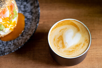 Heart latte art coffee scene with a bagel with egg, avocado, and fish on a wooden table. Perfect breakfast or brunch