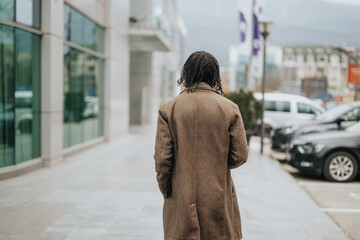 Obraz premium Rear view of a trendy individual with dreadlocks wearing a winter coat, strolling on a city sidewalk, exuding independence and style.