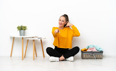 Young caucasian woman folding clothes sitting on the floor isolated on white background making phone gesture and pointing front