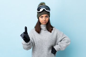 Mixed race skier girl with snowboarding glasses over isolated blue background counting one with...