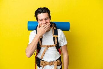 Young mountaineer Russian man with a big backpack isolated on yellow background happy and smiling covering mouth with hand