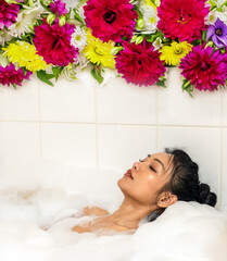 A young woman lies in a bathtub with foam, painting of flowers above her head