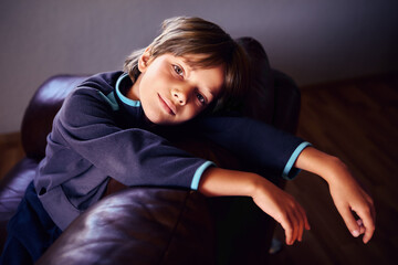 Obraz premium Sad male child is deprived of friends and gadgets as punishment for pranks, he is bored in dimly lit room alone, sitting on brown leather sofa.