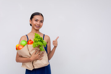 Positive young Asian woman holding a paper bag full of fresh groceries and showing thumbs up on a...