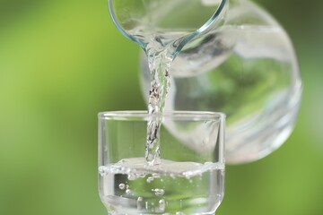 Pouring fresh water from jug into glass against blurred green background, closeup
