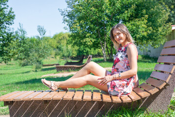 a beautiful slender girl in a bright elegant dress sits on a sun lounger in the park against the blue sky.