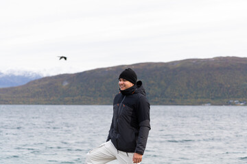 Fototapeta premium hombre latino con ropa de invierno con fondo desenfocado de fiordo noruego y ave volando