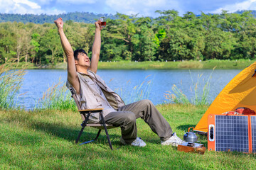 Asian male man sitting on chair, drinking coffee, feeling relax and raising hands, with camping tent beside lake and mountain, with mobile solar cell.