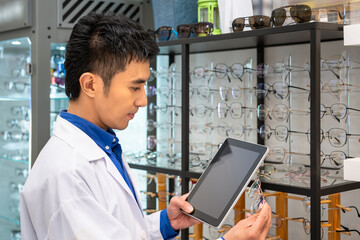 Portrait of young Asian male man optician in optical shop store, holding digital tablet. Eyecare...
