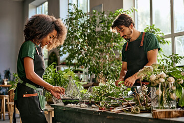 Two entrepreneurs work in a plant shop, transplant flower pots, make florariums. Beautiful african-american woman and a caucasian men in aprons work at a table with plants. - obrazy, fototapety, plakaty