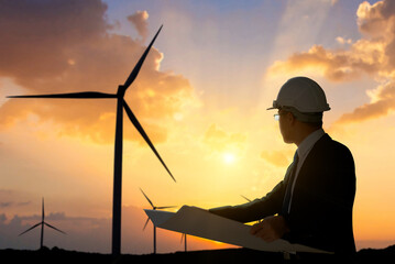 .An engineer in hard hat reviews plans against a backdrop of wind turbines and a dramatic sunset.