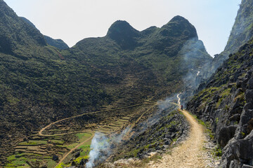 A winding mountain trail with smoke rising in a lush valley
