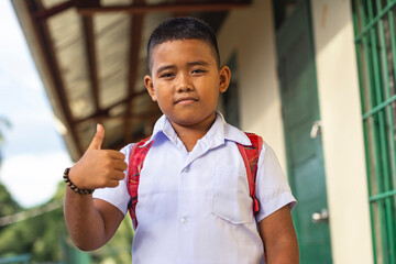 Confident young boy student with backpack outside classroom showing thumbs up, ready for school day. - obrazy, fototapety, plakaty