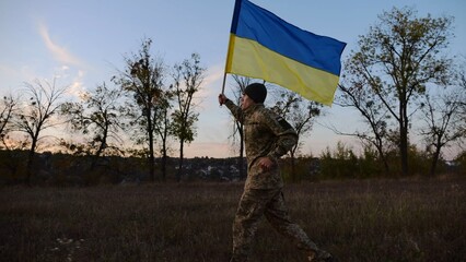 Soldier of ukrainian army running with raised blue-yellow banner on field at dusk. Young male...