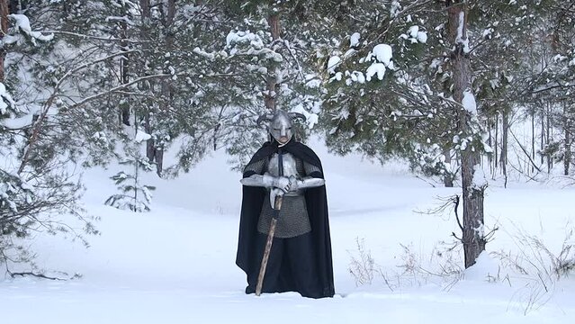 Portrait of a medieval fantasy warrior in a horned helmet, steel breastplate, chainmail with a two-handed ax in his hands, posing against the backdrop of a winter forest. Camera movement.