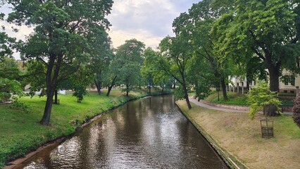 Along the banks of the canal, one of which is reinforced with timbers, there are paved walkways, grass, maples and linden trees. The water is rippling. Buildings stand nearby. Sunny summer weather