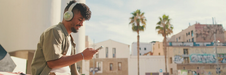 Young male student dressed in an olive color shirt sits outside next to the university, listens to music on headphones, selects tracks on his phone, Panorama