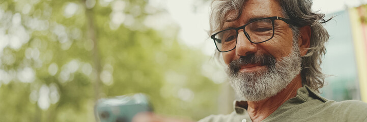 Panorama, Positive middle-aged man with gray hair and beard wearing casual clothes sits on bench and uses mobile phone. Mature gentleman in eyeglasses writes message on smartphone - Powered by Adobe