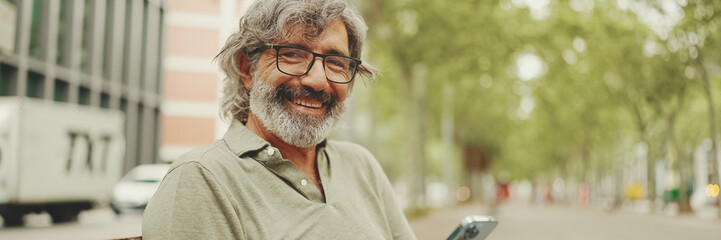 Panorama of friendly middle-aged man with gray hair and beard wearing casual clothes sits on bench....