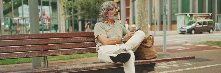 Panorama of positive middle-aged man with gray hair and beard wearing casual clothes sits on bench...