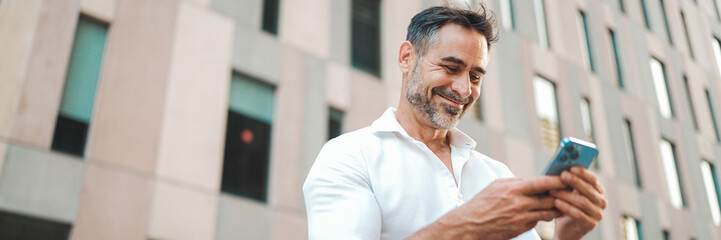 Mature businessman with neat beard uses mobile phone sits on bench in the financial district in the city, Panorama. Successful man scrolls through information on smartphone and smiles