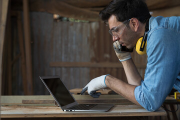 Male carpenter using laptop computer at woodwork workshop. Male joiner working with laptop computer...