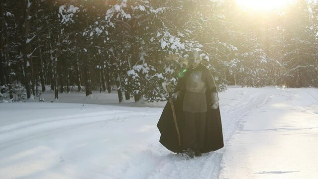 A medieval fantasy warrior in a horned helmet, a steel breastplate and chain mail with a two-handed ax in his hands walks through a winter forest against the backdrop of sunset. Selective focus
