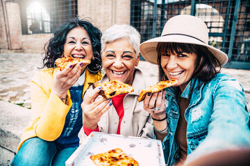 Happy friends enjoying street food in the city center - Three senior women eating pizza slice...
