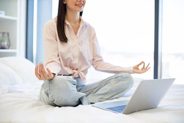 Poster Cropped view of woman enjoying practicing yoga during break with modern laptop at home. Peaceful lady sitting with closed eyes on bed on lotus position during online video class using modern laptop. © sofiko14