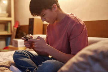 Side view portrait of young teenage boy playing mobile games at night sitting on bed in dim light