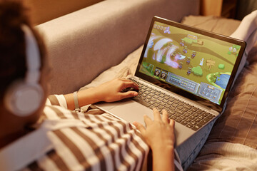 Close up of unrecognizable teenager playing online video games via laptop while lying on bed at...