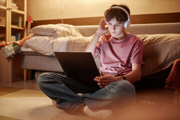 Fototapeta na wymiar Full length portrait of young teenage boy using laptop while sitting on floor at night with legs crossed and wearing wireless headphones