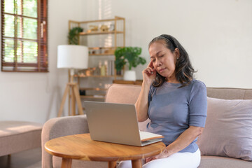 Older asian woman appears stressed and fatigued while working on a laptop from her home.