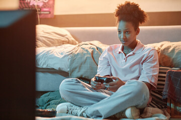 Portrait of teenage girl holding gamepad and playing videogames sitting on floor at night, copy...