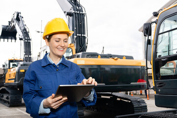 Woman engineer in a helmet with a digital tablet stands next to construction excavators