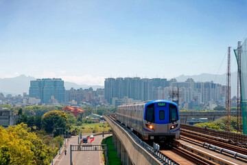 Fototapeta premium Scenic view of a metro train traveling on elevated rails of Taoyuan Airport MRT System and residential buildings clustering in background under blue clear sky in Zhongli, Taoyuan City, Taiwan, Asia