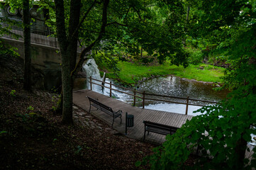 Along the trail is a sitting bench looking out at waterfall and park in a shaded area in summertime. Peaceful place to solitude and relax. Vecais parks, Smiltene, Latvia.