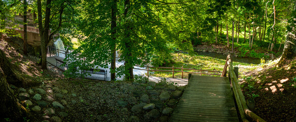 Along the trail is a sitting bench looking out at waterfall and park in a shaded area in summertime. Peaceful place to solitude and relax. Vecais parks, Smiltene, Latvia.