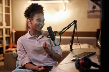 Portrait of smiling Black teenage girl speaking to microphone while live streaming videogames or...