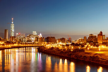 Obraz premium Riverside urban skyline of Taipei, the vibrant capital city of Taiwan, with landmark 101 Tower among modern skyscrapers in Xinyi District and city lights reflected on Keelung River in evening twilight
