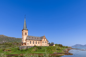 Fototapeta na wymiar Vagan Kirke, a striking historical church in Lofoten, Norway, stands proudly by the waterside, its spire reaching into the clear sky amid a serene natural setting