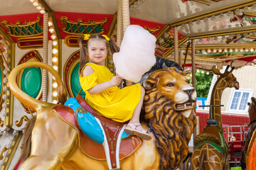 Fototapeta na wymiar Adorable little girl in summer yellow dress at amusement park having a ride on the merry-go-round and eating cotton candy