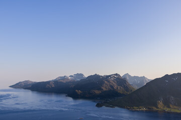 Evening descends on the Lofoten Islands, Norway, with the setting sun casting a warm glow on the imposing mountain range and the tranquil Norwegian Sea hugging the coastline