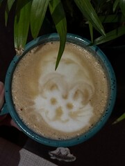 Cute kitten on milk foam. Cat latte art. Latte art for children. Foreground plant leaves and a cup of coffee. 