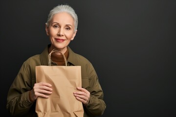 Fototapeta premium Middle age business concept. Smiling mature woman in jacket holding paper bag mockup, on studio background with copy space