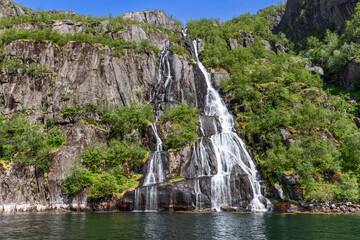 Fototapeta na wymiar A striking waterfall splits into silvery threads down a moss-draped cliff into the emerald waters of Trollfjorden in Lofoten, Norway