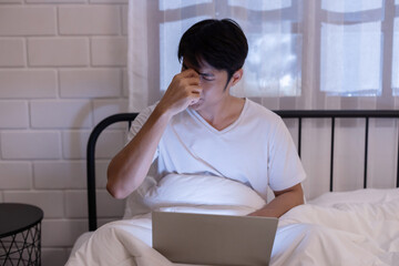 Sleepy asian man suffer from working overtime in front of laptop on his bed at night